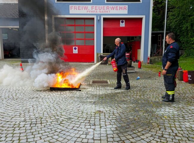 Ein Mann löscht vor dem Feuerwehrhaus unter Aufsicht mit einem Feuerlöscher ein Feuer in einer Brandtasse