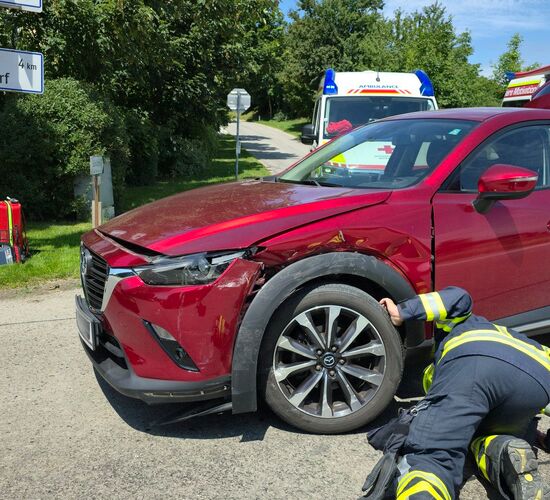Ein roter PKW auf der Kreuzung mit Kollisionsschaden auf der linken Vorderseite, im Hintergrund 2 Rettungsfahrzeuge