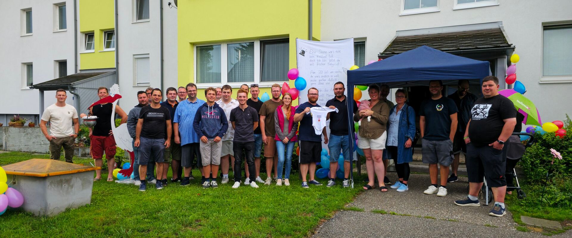 Foto: Alle Gratulanten vor dem aufgestellten Storch, Kommandant und Papa halten einen der überreichten Bodys für die Kleine Annika