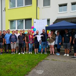 Foto: Alle Gratulanten vor dem aufgestellten Storch, Kommandant und Papa halten einen der überreichten Bodys für die Kleine Annika