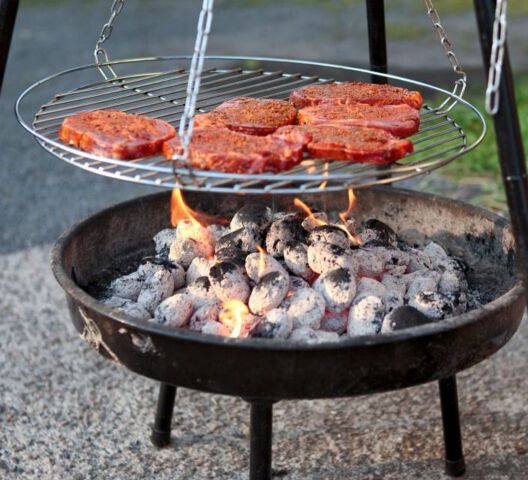 Ein dreibeiniger Griller, unten die Schale gefüllt mit heißer Kohle, darüber hängt ein Grillrost mit Fleisch